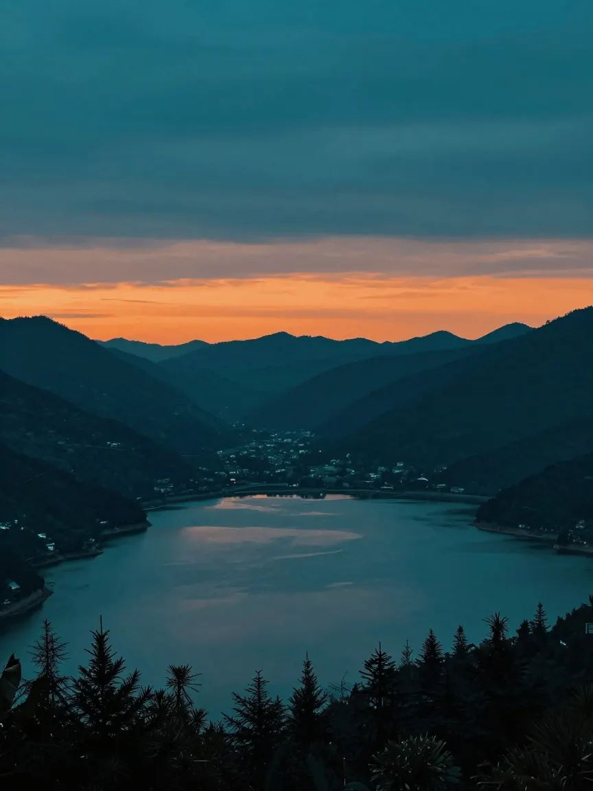 Dusk view over a lake ringed by dark forested mountains, orange band of sky above the ridgeline, empty sky across the top of the frame